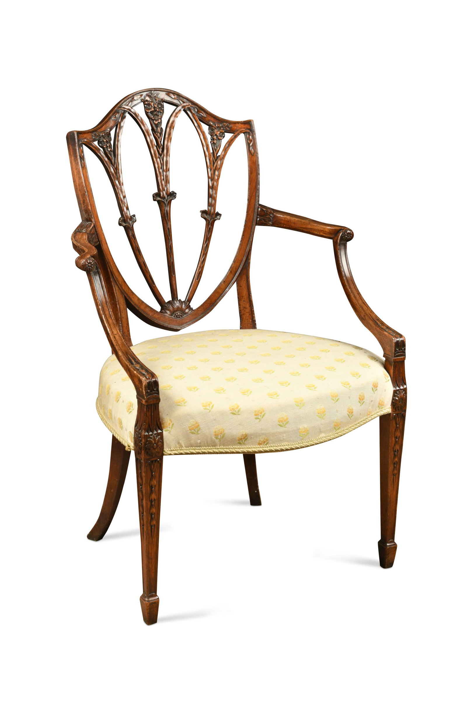 A George III mahogany Hepplewhite style elbow chair,: the shield shape back with carved ribbon and flower head decoration between scrolling arms raised on square tapered front legs with bellflower detail on spade feet, the stuff over seat upholstered in 