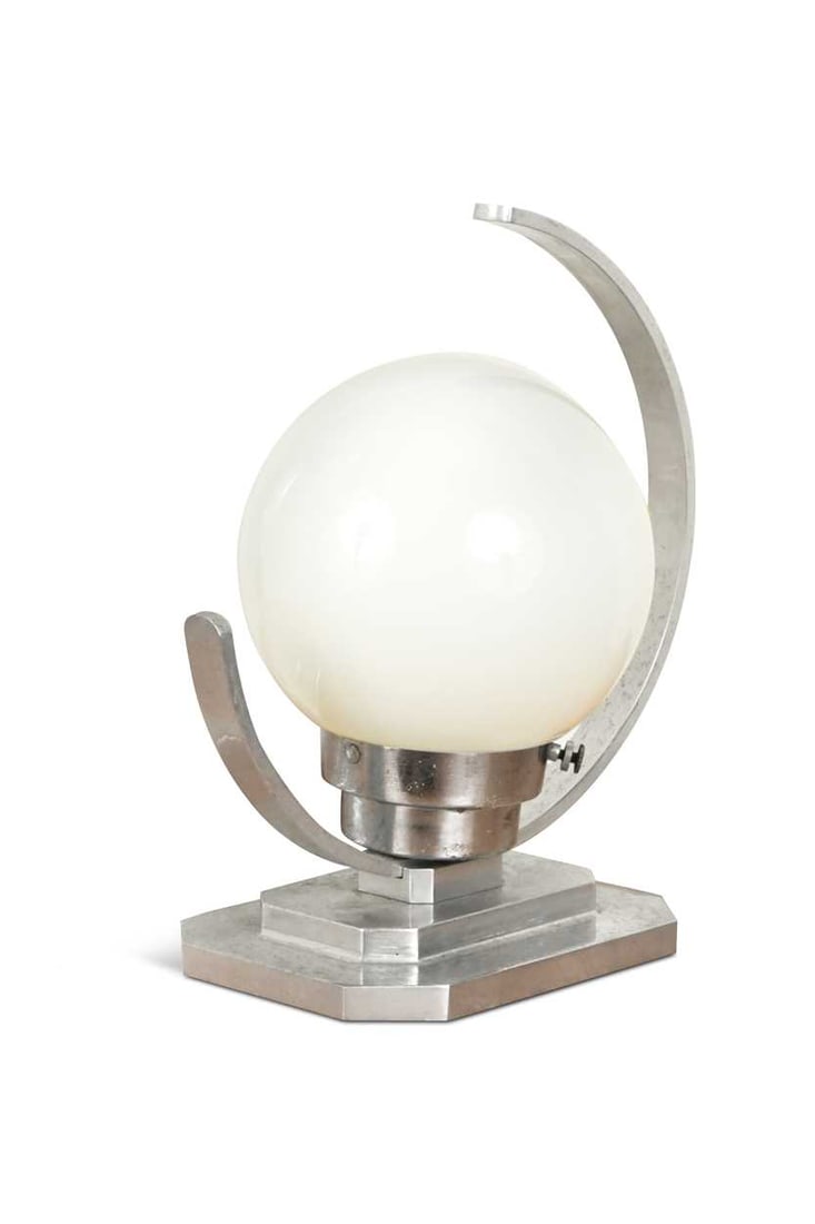 An Art Deco aluminium table lamp, the milk glass globe within a canted C-shape frame and raised on a: An Art Deco aluminium table lamp, the milk glass globe within a canted C-shape frame and raised on a stepped base 28cm high