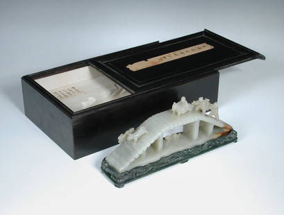 An ebony boxed nephrite jade bridge scene on spinach: An ebony boxed nephrite jade bridge scene on spinach green stand, the pale grey green stone carved with a horseman and two carriers following a dog across the bridge with two ducks swimming below, the