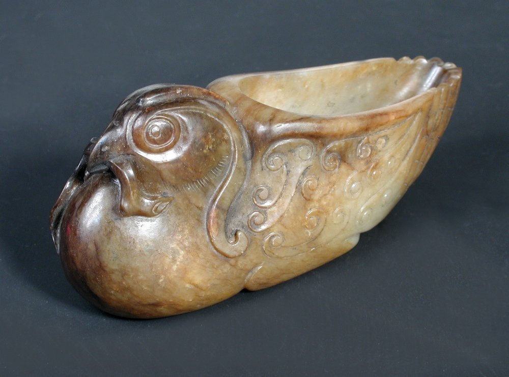 A late 18th/early 19th century jade libation cup,: A late 18th/early 19th century jade libation cup, the pale green stone with areas of brown crust, carved as a bird with lingzhih fungus in its beak, its feathers forming tight archaic scrolls, 10.5cm