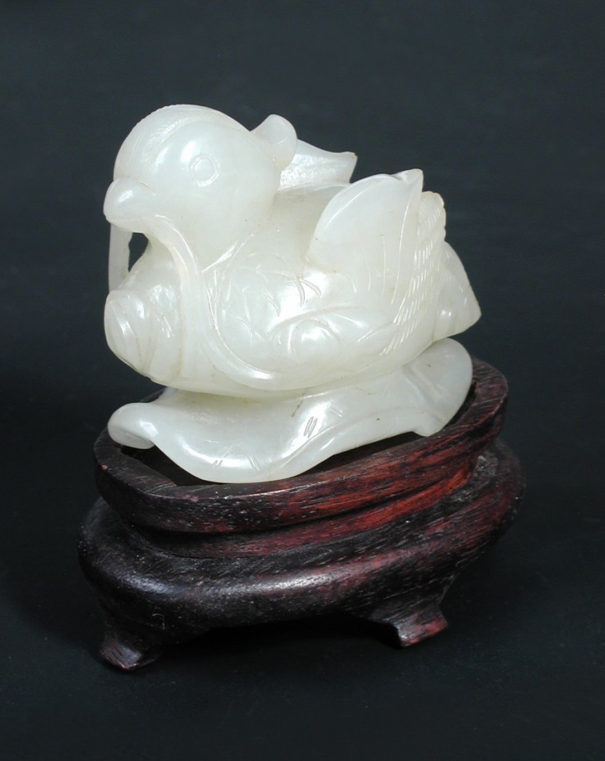 A white jade mandarin duck, possibly late Ming,: A white jade mandarin duck, possibly late Ming, the bird with upswept wings with lotus in its beak, the supporting leaf pierced with four holes, 5cm (2 in ) wide, with a wood stand (2)