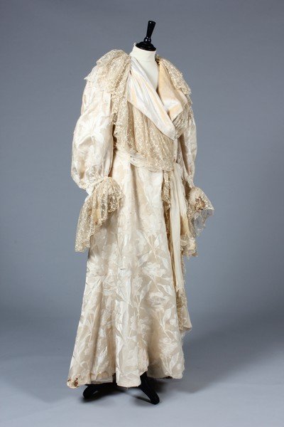 A poor condition Charles Frederick Worth ivory damask: A poor condition Charles Frederick Worth ivory damask deshabille robe, circa 1900, trimmed with cascades of embroidered lace, original waist label applied to the neck, and re-lined in velvet in the 19