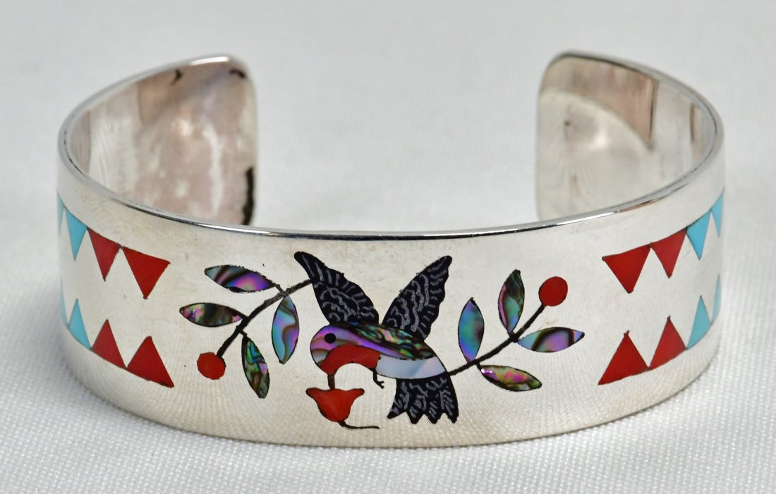 Dennis Edaakie Sterling Silver Hummingbird Inlay Cuff: Measuring 3/4 inch wide all around. This piece has Inlay of a Hummingbird in flight across the top. The inlay is of Coral, Turquoise, Mother of Pearl and Abalone. Inside measures 5.25 inches plus a ga