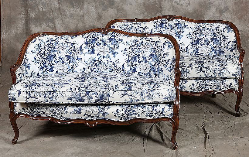 Pair of LVX Style Carved Settees: Scrolled and carved walnut bow front frames in blue and white floral upholstery. Age: Mid 20th Century Condition: Very Good: Minimal wear and dings to the frames. Some finish loss. Clean serviceable u