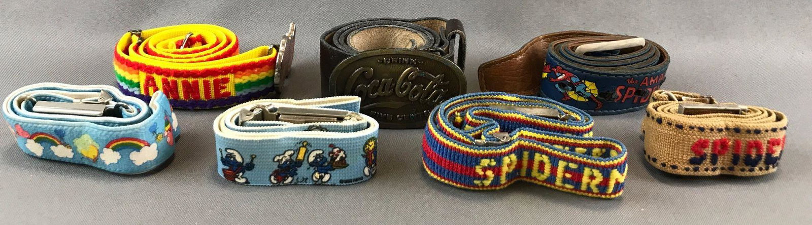 Group of 7 Vintage Leather and Elastic Licensed: Annie, Coca-Cola, Spider-Man, Care Bears, The Smurfs. Largest is 1.5x31 inches.