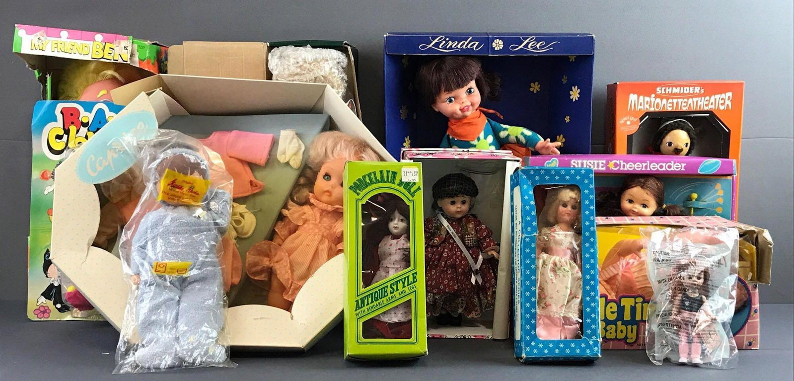 Group of 13 assorted dolls: Includes porcelain, cloth, plastic and more. Includes sleep eye dolls, largest measures 15.5x11.5x3 inches. In original packaging, some shelf wear, rips and cellophane detached and missing.