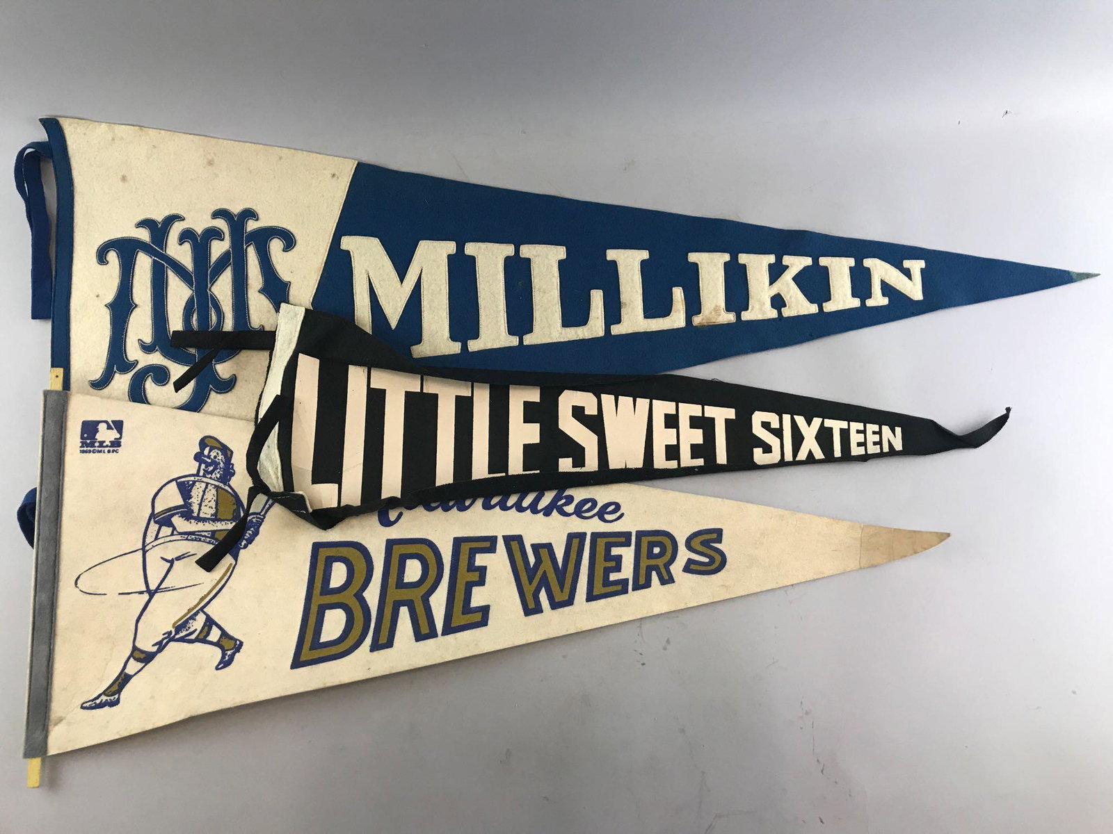 Group of 3 vintage felt pennants: Largest is 14x36 inches. Millikin is embroidered, Milwaukee Brewers and Little Sweet Sixteen are printed. Some staining/discoloration