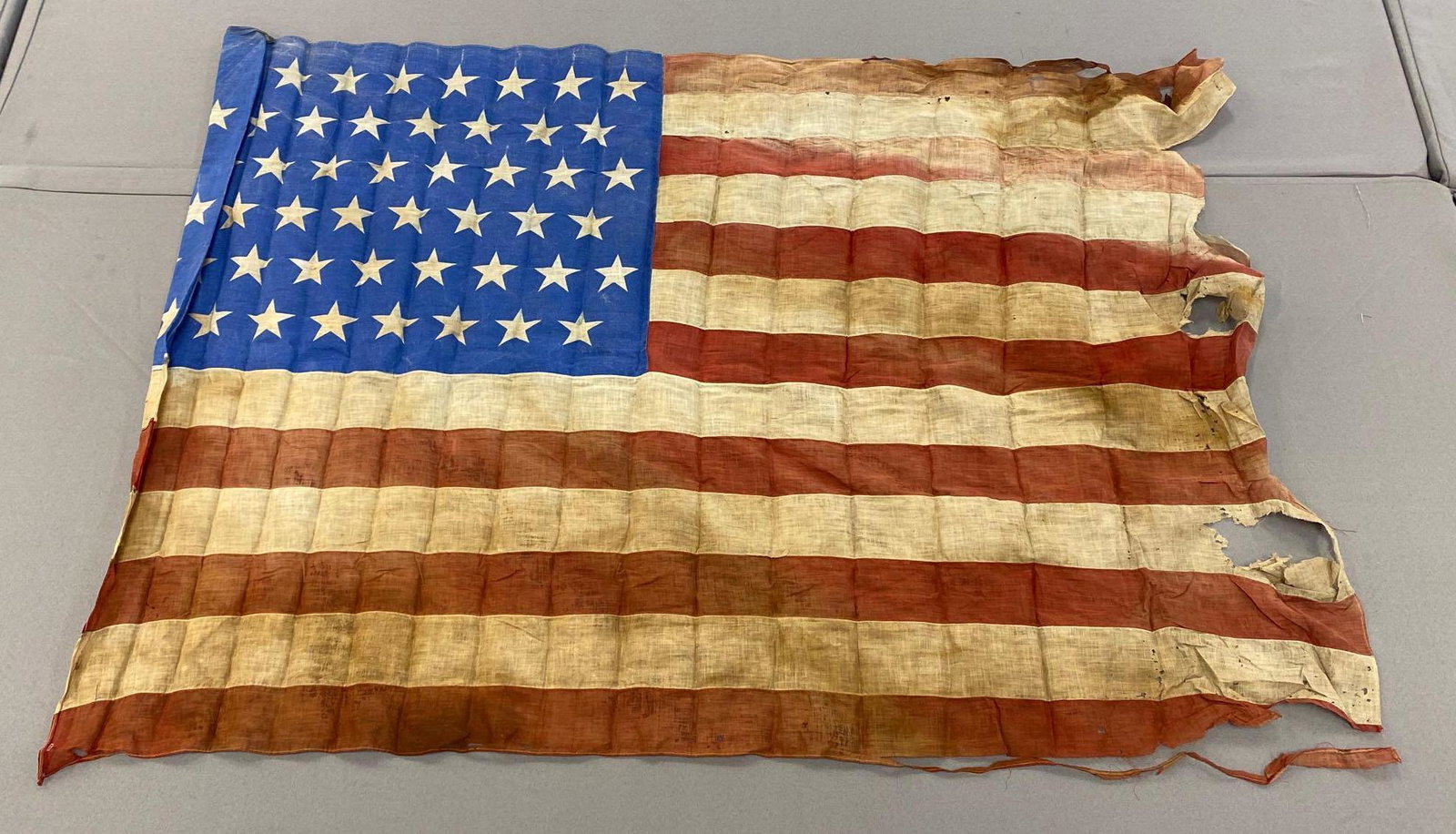 48-Star American Flag: Weather-worn flag. Measures 53x37inches. Right side is in pieces. Machine stitched, several stains and tears.