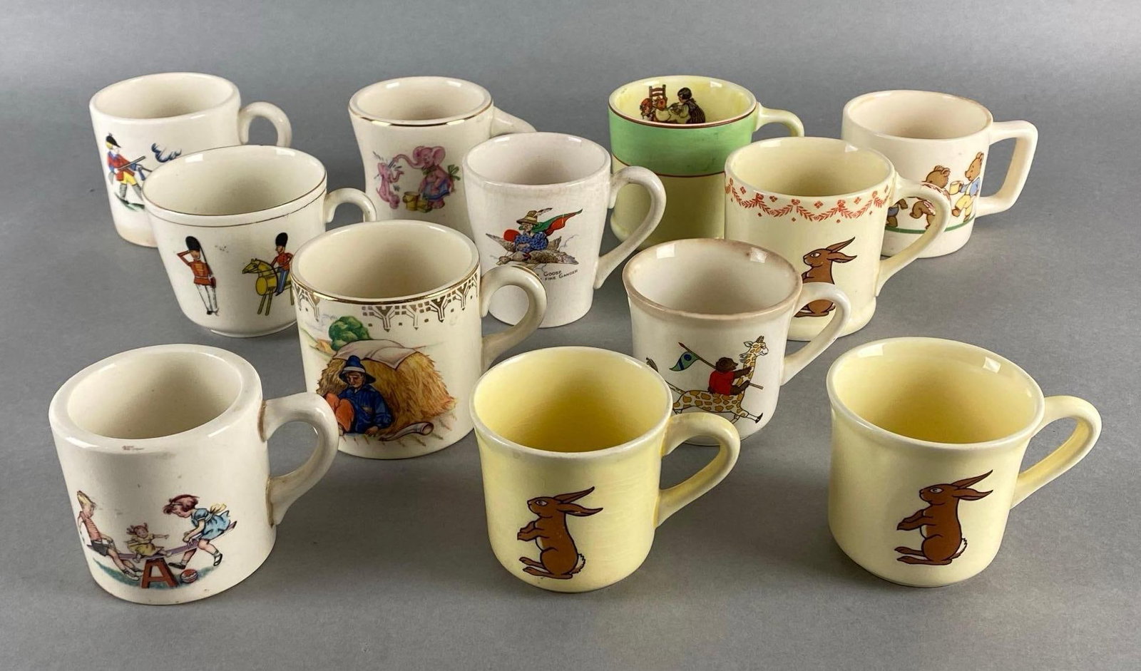 Group of 12 Porcelain Childrens Nursery Rhyme Mugs: Includes Mother Goose, Little Boy Blue, Toy Soldiers, Rabbits, and more, Some cracks and chips throughout, Largest measures 3 x 4 1/4 x 3 inches.