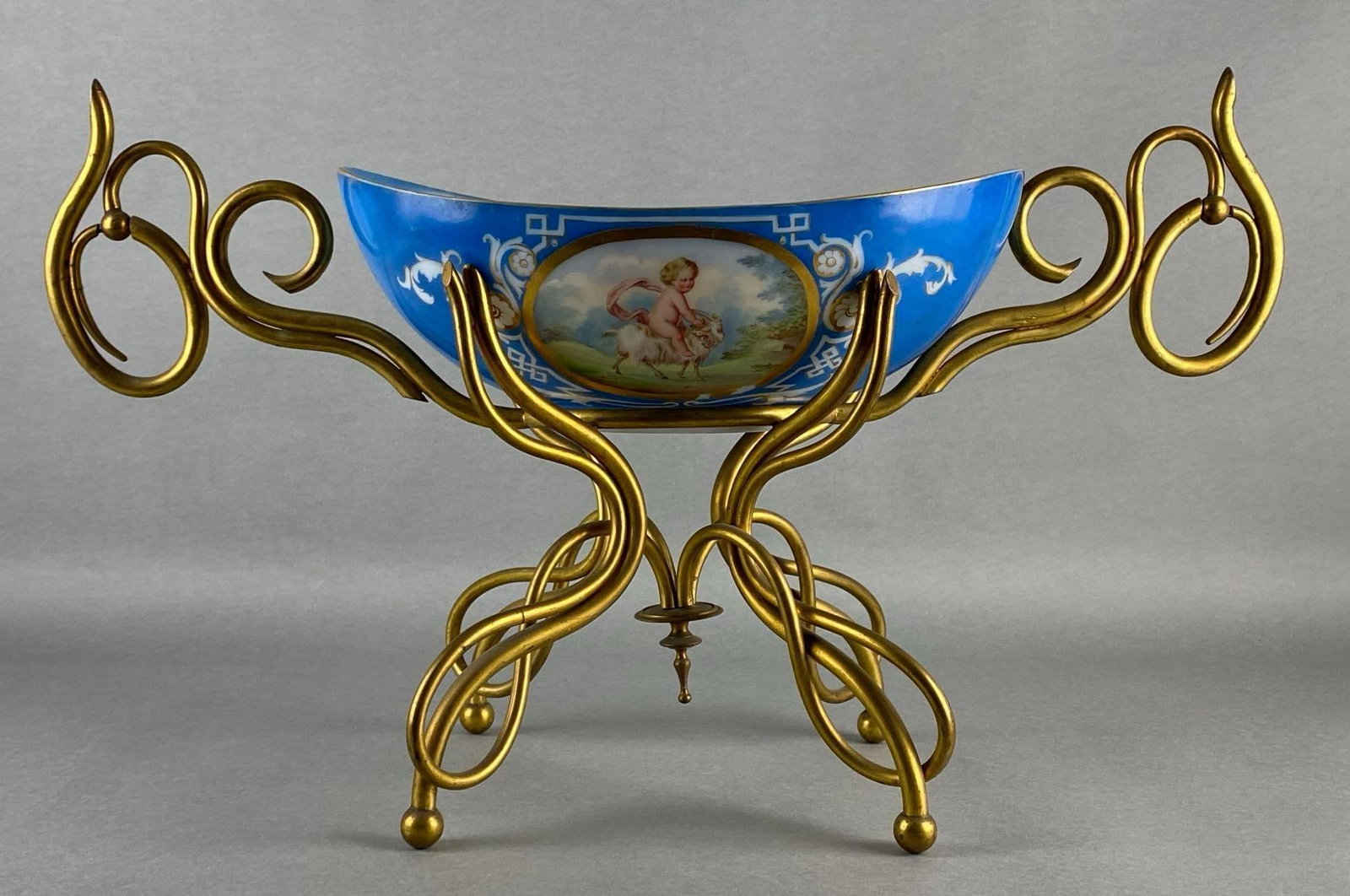 Baccarat Porcelain Figural Centerpiece Bowl on Ornate Brass Stand: Painted child riding ram, Painted gold gilt accents, Handles not fully attached, One handle missing prong to hold in place, Measures 14 3/4 x 25 x 10 inches.