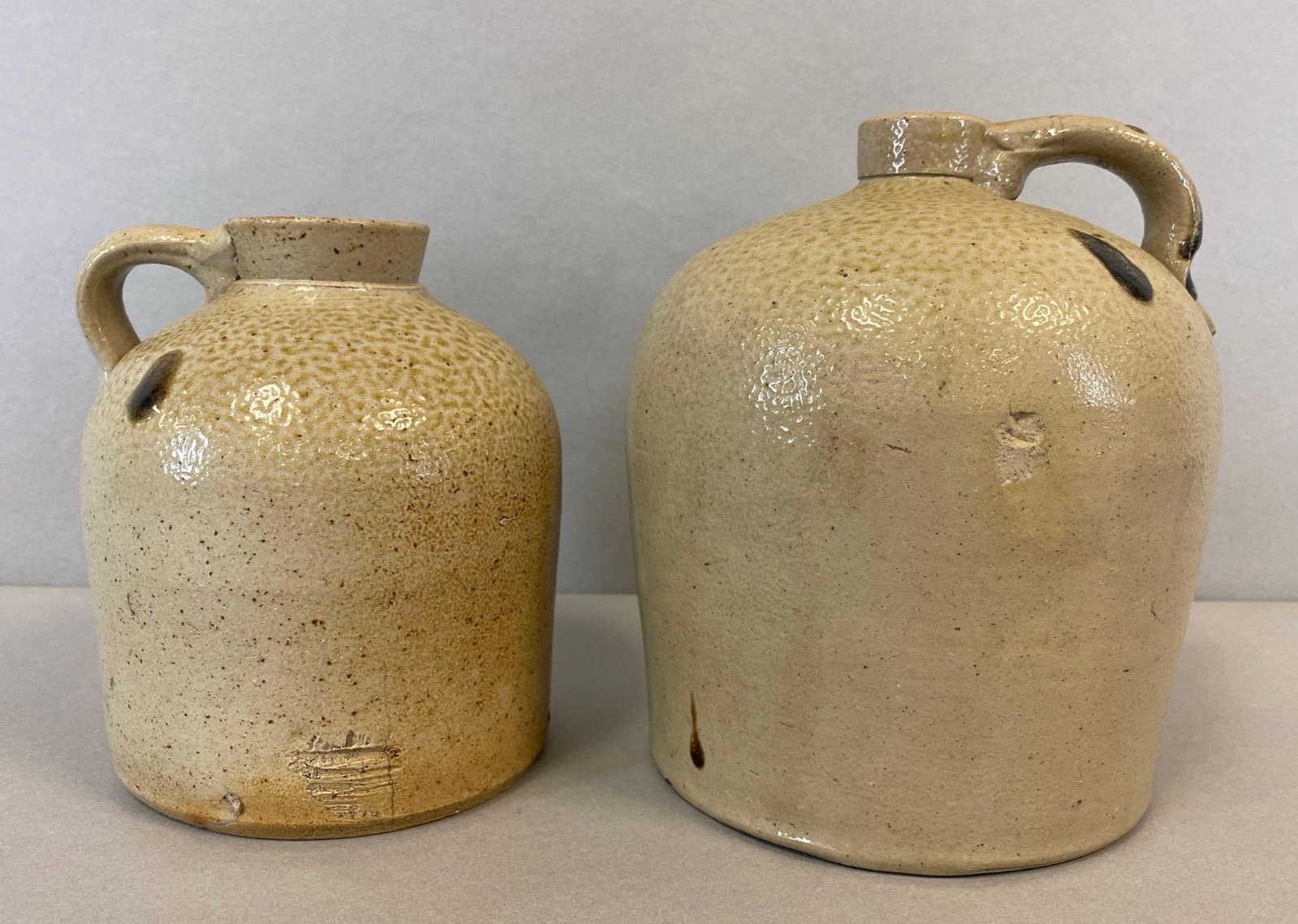 Group of 2 Antique Stoneware Salt Glaze Jugs: Larger jug is a Gallon. Smaller jug is a half gallon. Larger jug has a crack on the handle and chip. Both jugs have turkey droppings. Largest measures 9 x 7 x 7 inches.