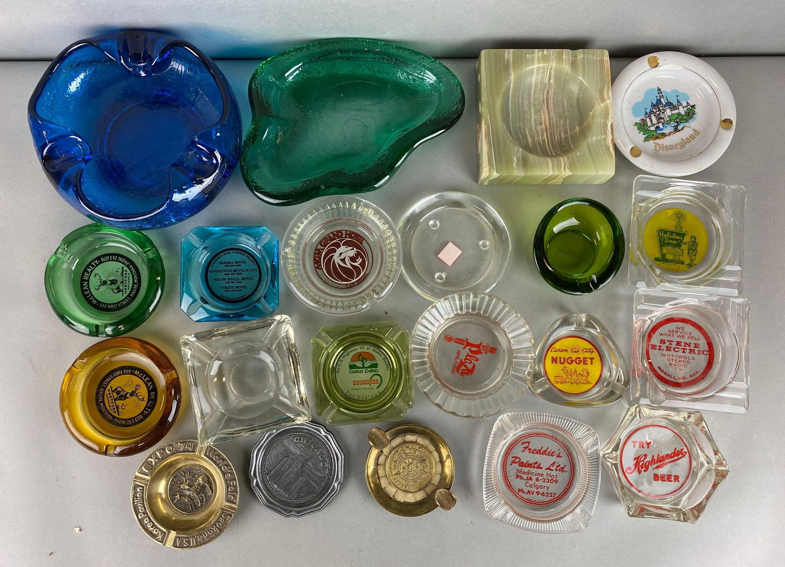 Group of 20 Vintage Ashtrays and More: Includes a cigar rest, Disneyland, world fair 1974 and more. Largest measures 3 x 7 x 7 inches.
