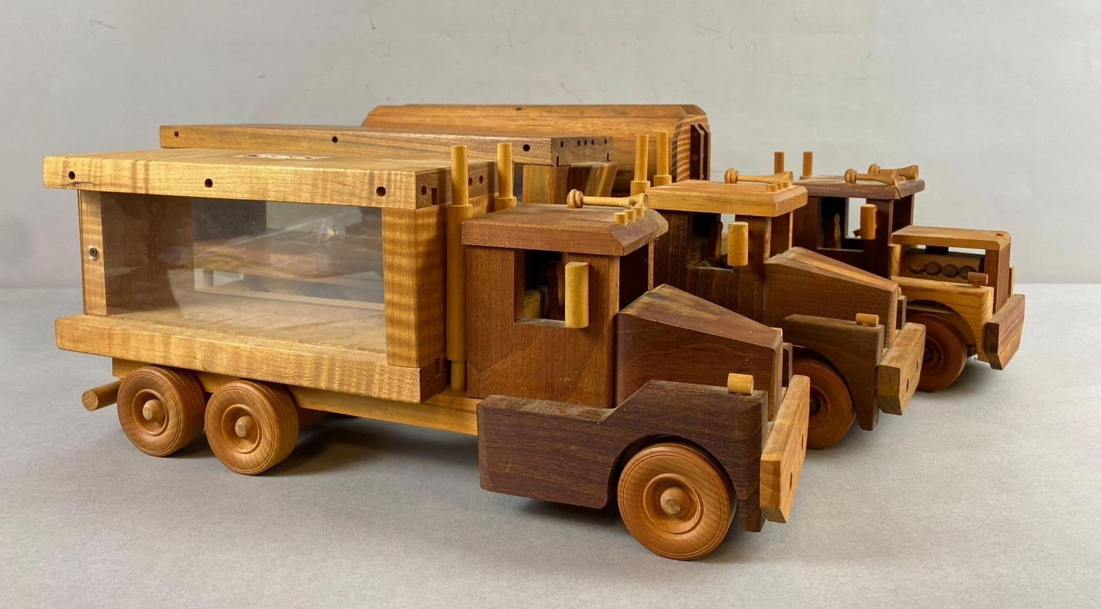 Group of 3 Handmade Cedar Wood Truck and Trailer Coin Banks: Little missing pieces throughout. Trailers act as coin banks and can stand on own. Largest truck and trailer measures 21 inches in length. One truck cab is loose from body.