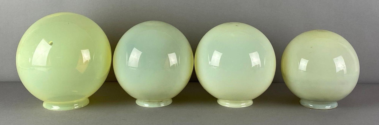 Group of 4 Vaseline Glass Globe Lamp Shades: Bruise shown on largest, fleabites in fitters, largest measures 8 inches H x 7 1/4 inches W with a 3 1/2 inches Fitter