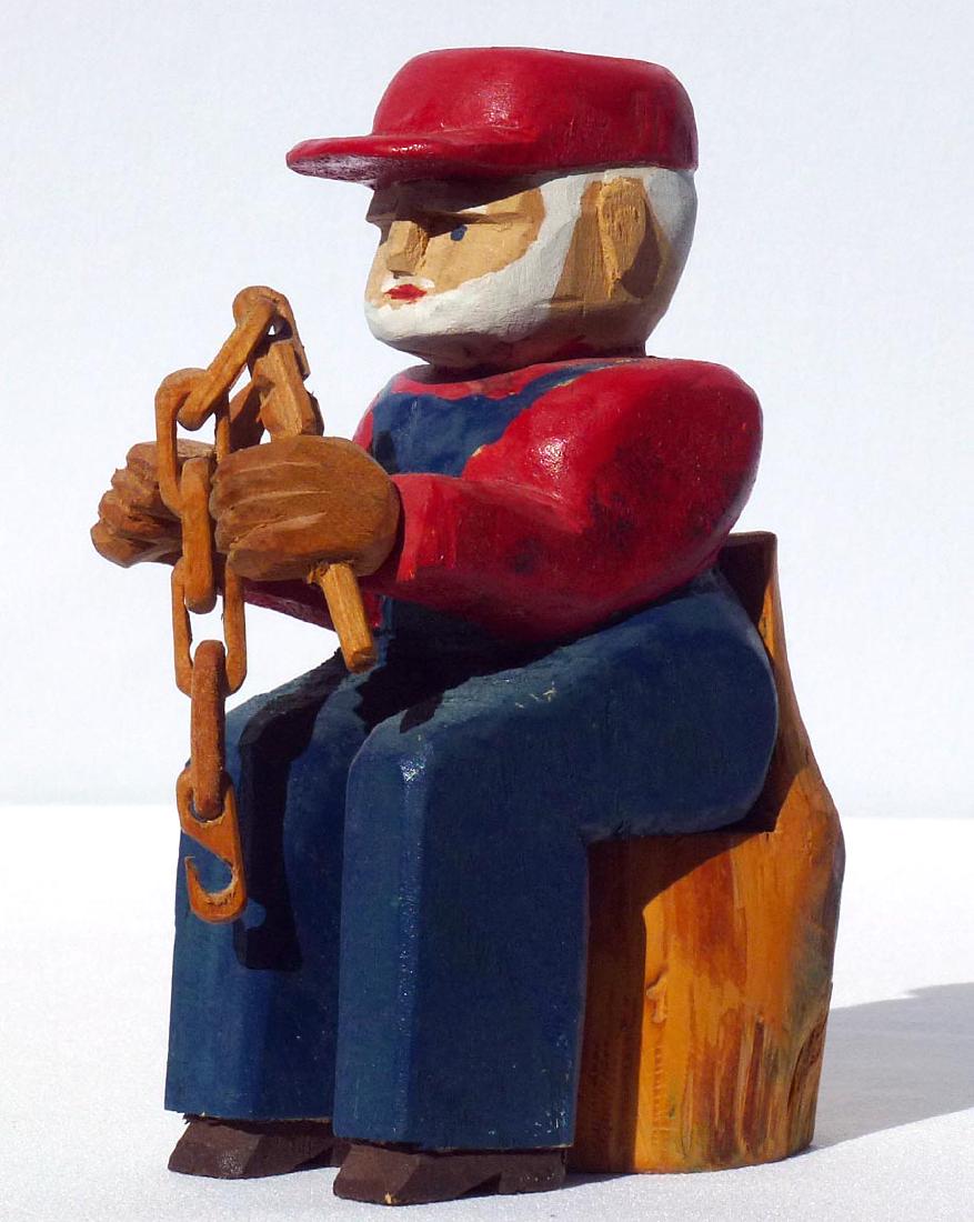Carving of a man who is carving a whimsy chain: This is a wonderful carving of a man who is carving a whimsy chain. The man has a beard, and is wearing painted overalls with a red shirt and cap. He is holding a knife in one hand. The whimsy in prog