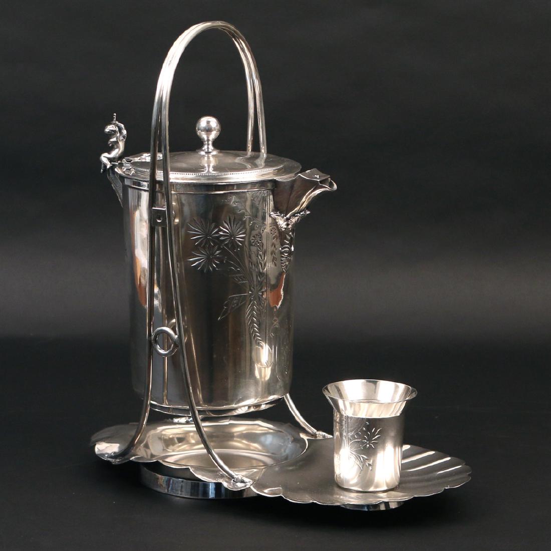 SILVER-PLATED PITCHER-ON-CRADLE: by Rogers, Smith, & Co., Connecticut, patented March 26, 1872, cylindrical lined pitcher with incised decoration and putti-form hinge, scallop-shaped cradle with matching cup, h. 16-1/2 w. 15 d. 7-1/2