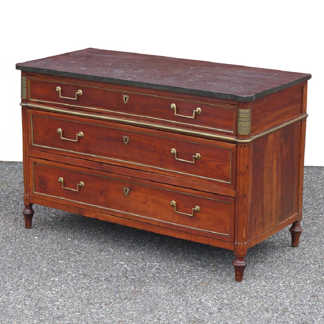 LOUIS XVI FRUITWOOD COMMODE: late 18th Century, with a black marble top containing ammonites with rounded outset corners over conforming reeded pilasters headed by brass mounts with classical bands all flanking three brass-lined 