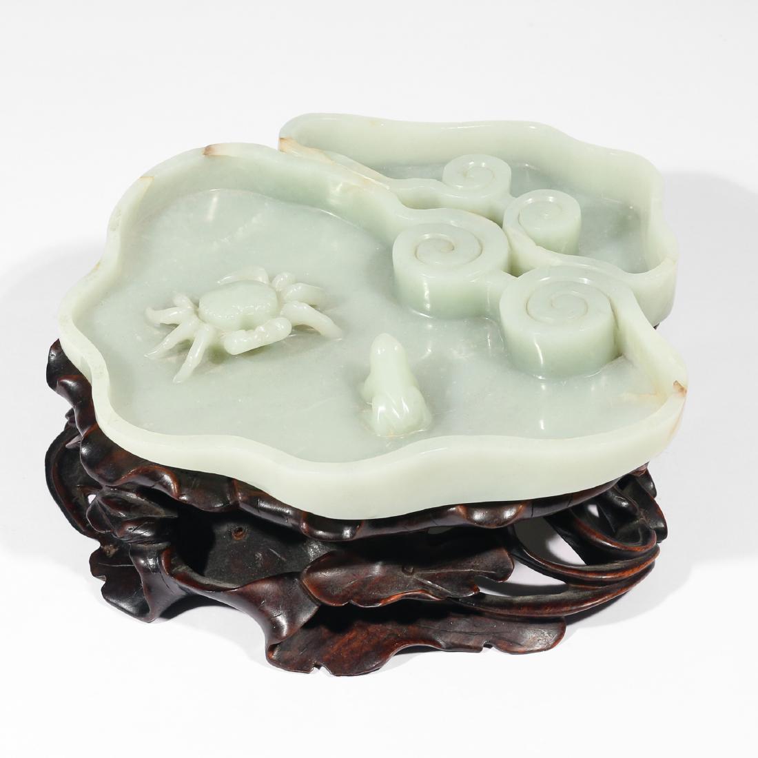 PALE GREEN JADE BRUSH WASHER: A carved jade low-form brush washer having two sections separated by a swirling cloud motif, the larger section with a crab and toad; resting on a conforming stand of carved wooden lily pads and vines