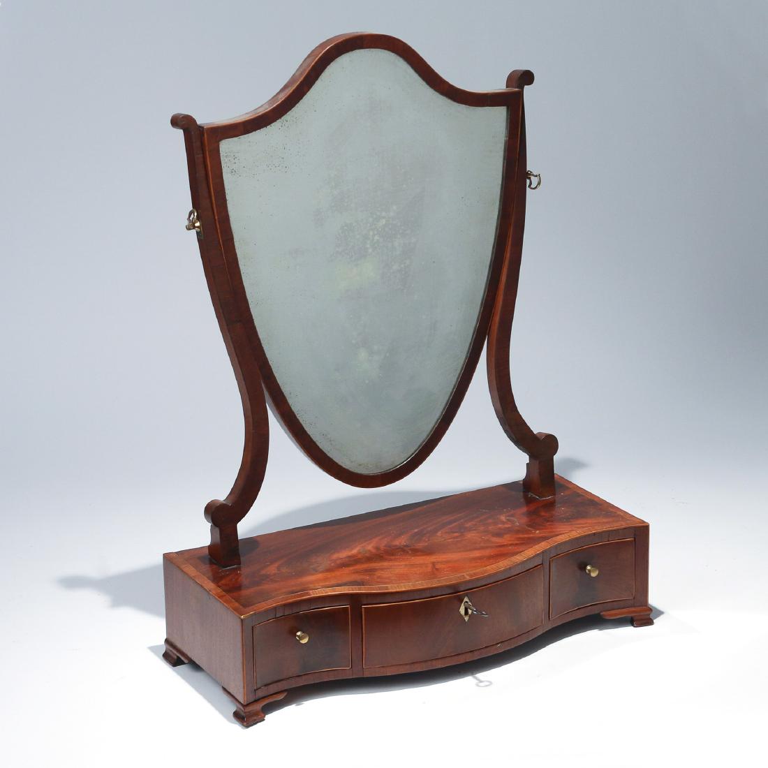 GEORGE III INLAID MAHOGANY DRESSING MIRROR: early 19th Century, the shield-shaped mirror above a serpentine bank of three drawers. h. 23-1/2 w. 17-1/2 d. 8 in. Provenance: Descent through the family of the late James Delihas, former curator at 