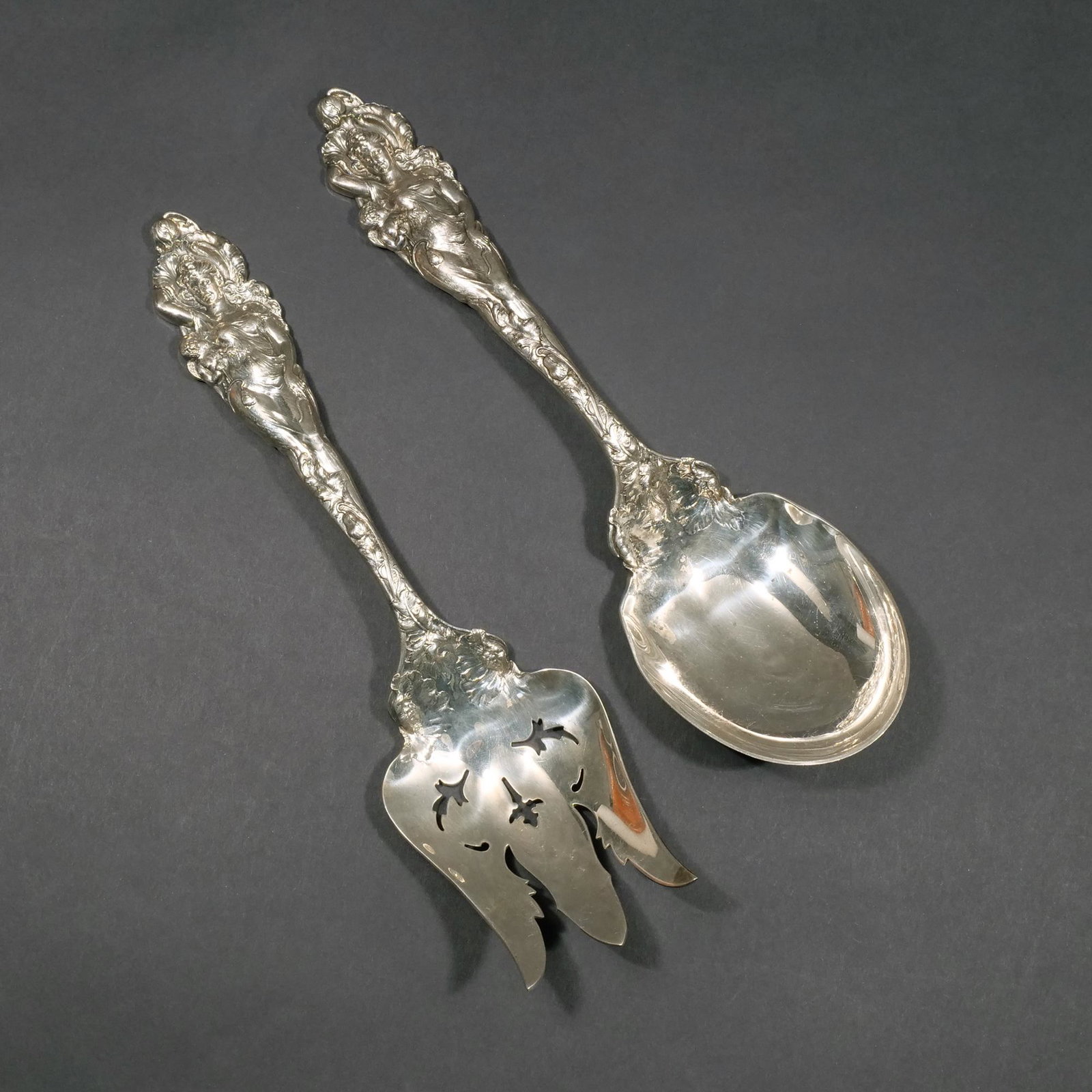 PAIR REED & BARTON "LOVE DISARMED" SERVERS: Decorated salad servers, 1899, showing female figures with putti in high relief among blooming flowers and vines, intricately engraved on both sides; 11 in., 15.7 ozt Provenance: Descended through a S