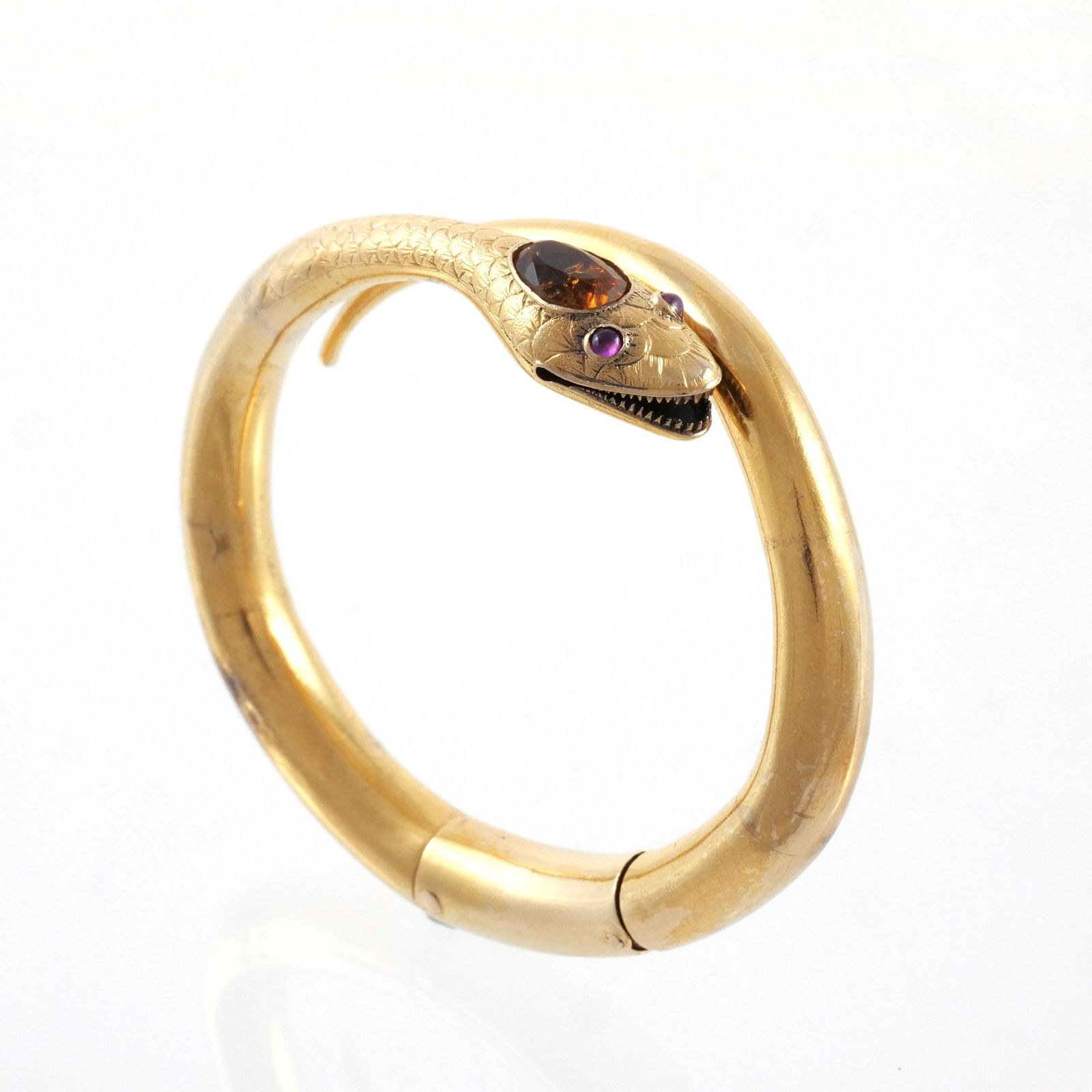 18K GOLD & GEMSTONE SNAKE HINGED BANGLE BRACELET: A coiled snake design with open mouth showing finely sharpened teeth, a scale-textured head and neck, cabochon eyes, and a faceted orange, pear-shaped gemstone on top of head, double-hinged, unmarked,