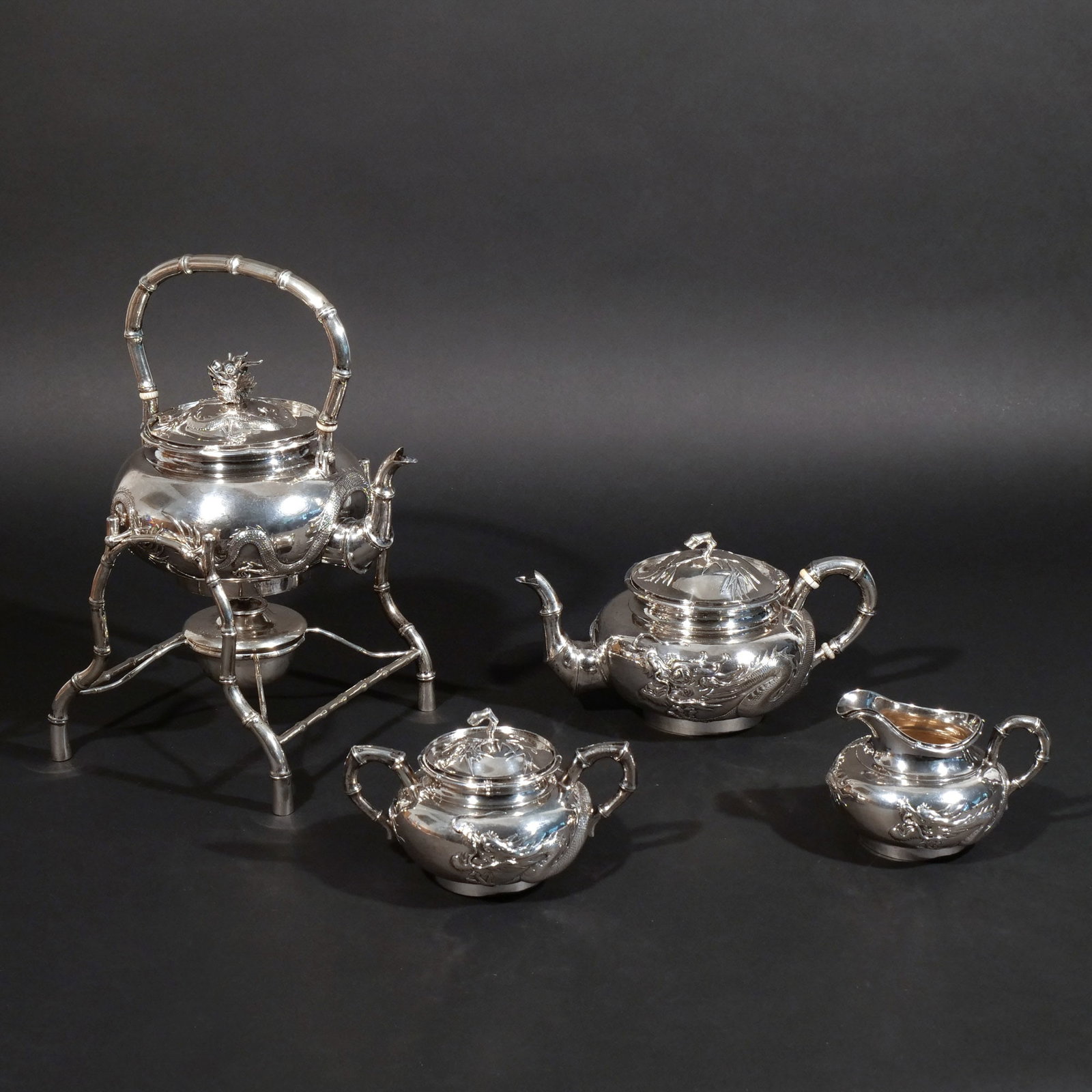 (4pc) WANG HING CHINESE EXPORT SILVER TEA SERVICE: Including a water kettle and stand, tea pot, creamer, and open sugar decorated with a sinuous dragon in relief; retailed by Wang Hing & Company, c. 1854-1925, Hong Kong, Canton, and Shanghai, marked W