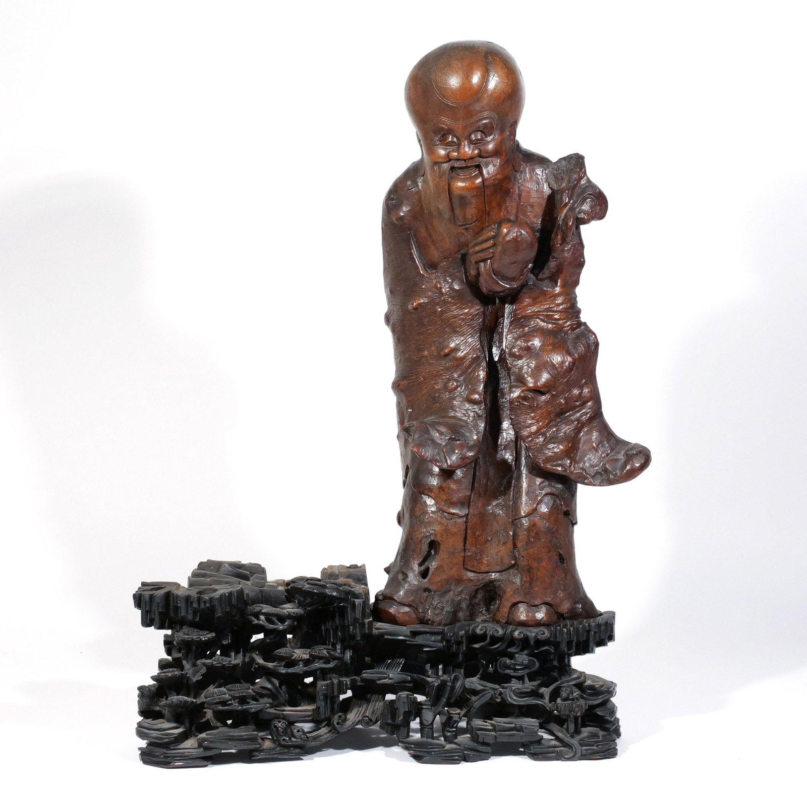 CHINESE ROOT CARVING of SHOU LOU: Late 19th / early 20th century, the god of Longevity carved from a large piece of burlwood, shown holding a peach, on a conforming carved wood base with rocks, lingzhi, and pine (symbols of longevity)