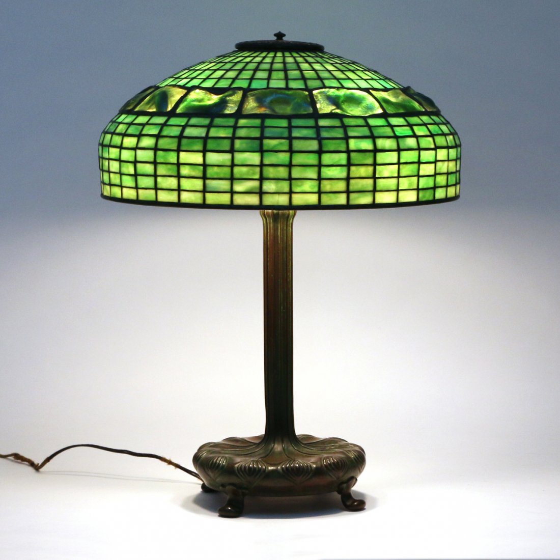 TIFFANY STUDIOS LEADED GLASS TURTLE TABLE LAMP: The domed shade composed of rectangular green tiles with a central band of ‘turtle-backs’ at the shoulder raised on a patinated bronze base of a circular lobed leaf design on four shaped feet, the