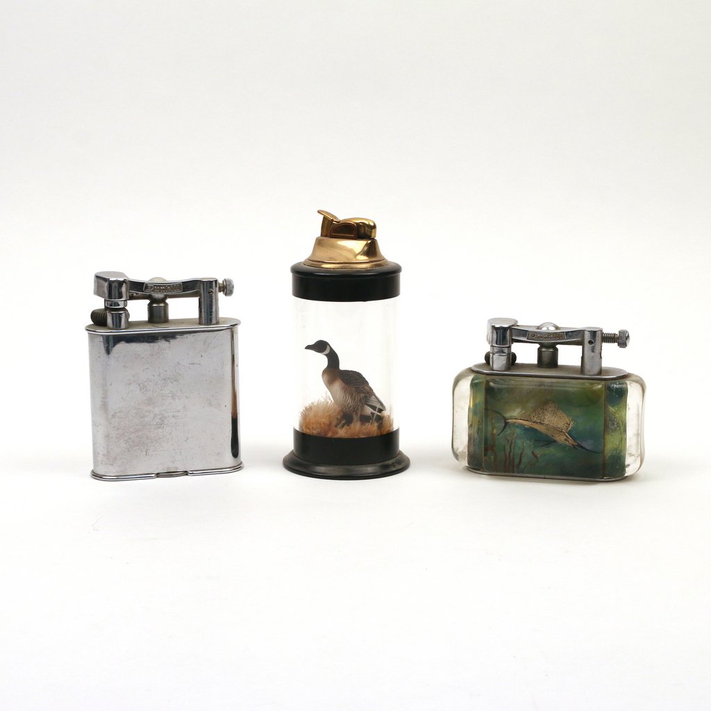 (3pc) LARGE DUNHILL & OTHER LIGHTERS: including a Dunhill lighter with an encased enameled scene of a marlin and a fish on the opposing side, h. 3 1/4 in.; a chrome Dunhill lighter, h. 4 1/4 in.; and a lighter with an encased model of a g