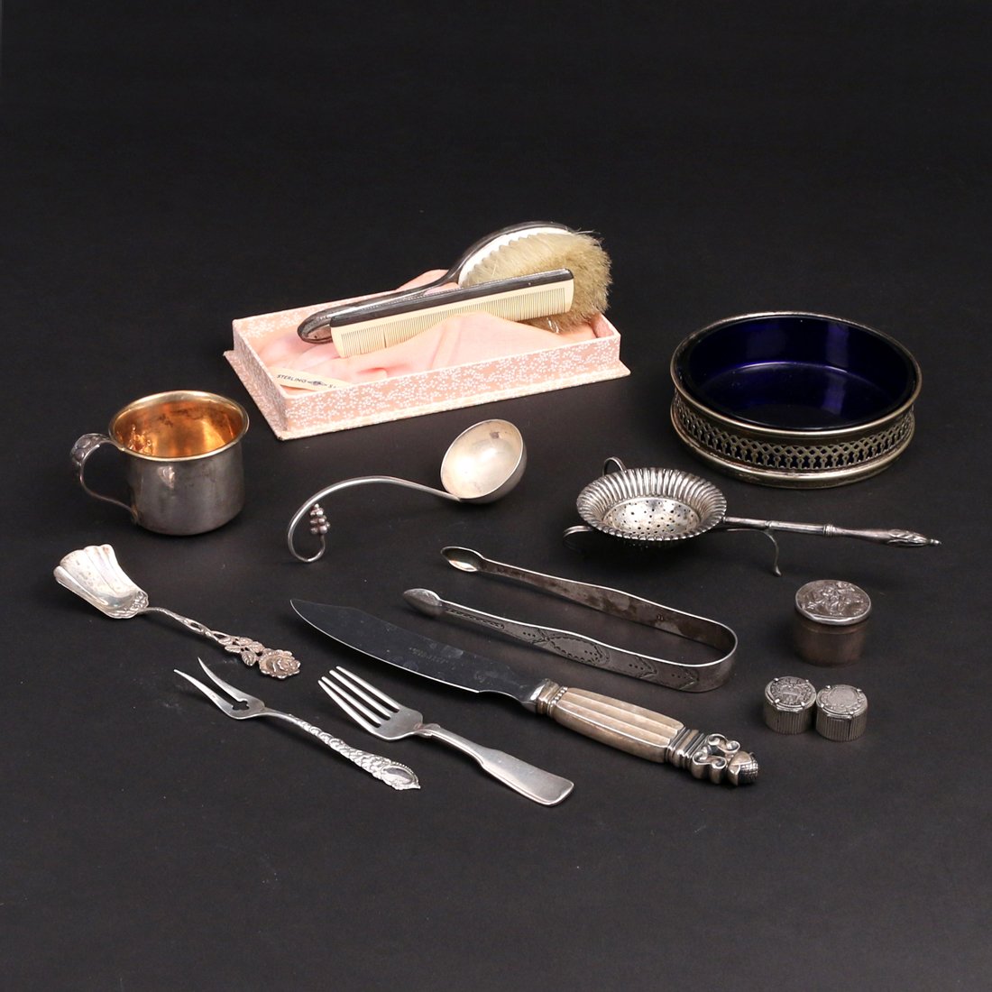 (12pc) MISC. SILVER ITEMS: Including a Georg Jensen Sterling fish knife, English ice tongs, a cream ladle, a wine coaster w/ cobalt blue insert (plated), tea strainer, two coin boxes, a baby cup, a baby fork, baby comb & brush,