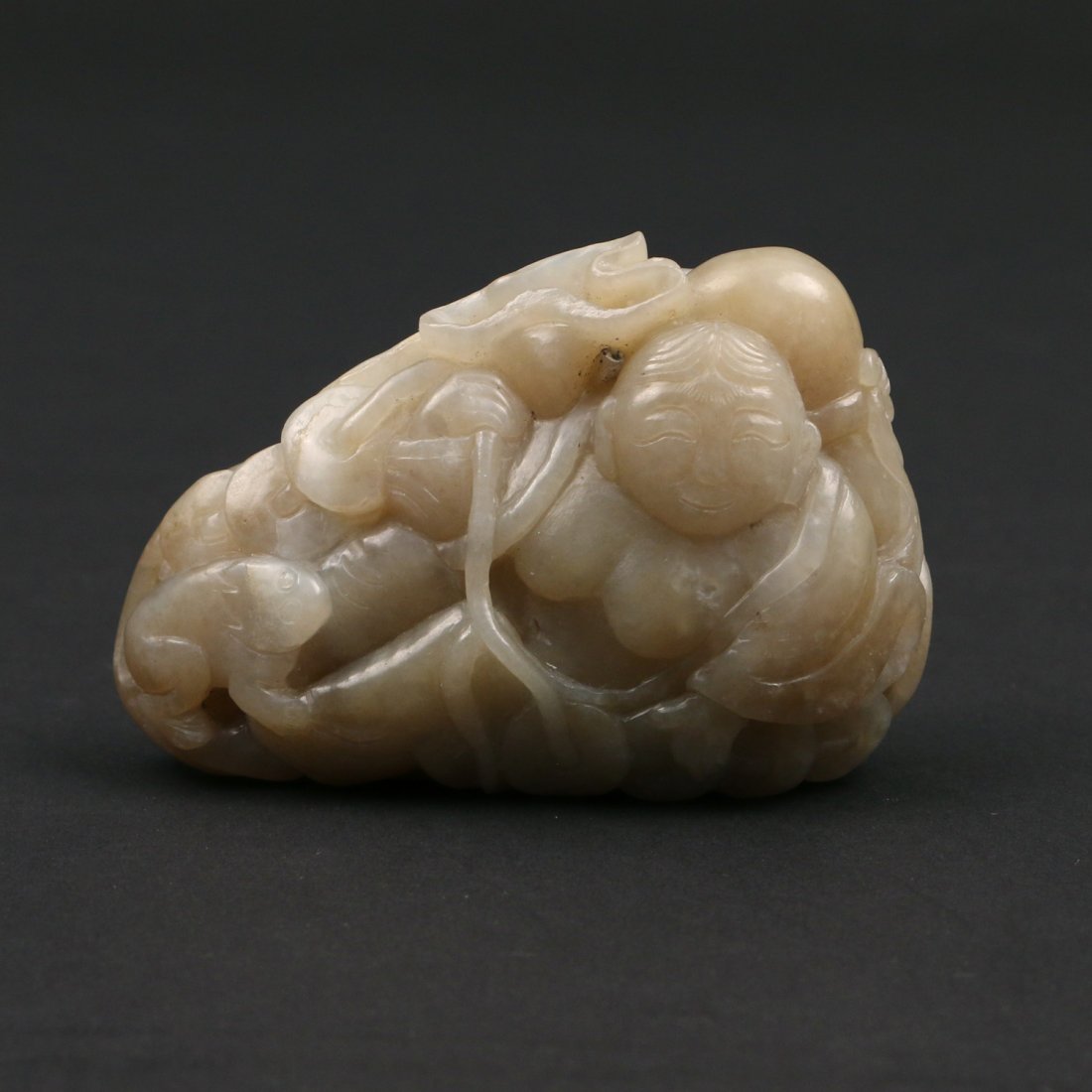 CHINESE JADE FIGURE OF LIU HAI WITH TOAD: The stone of a yellowish grey color with white and russet inclusions depicting Liu Hai reclining among on a large lotus leaf bearing his string of coins and with his toad by his feet, 2 x 3 x 1-1/8 in
