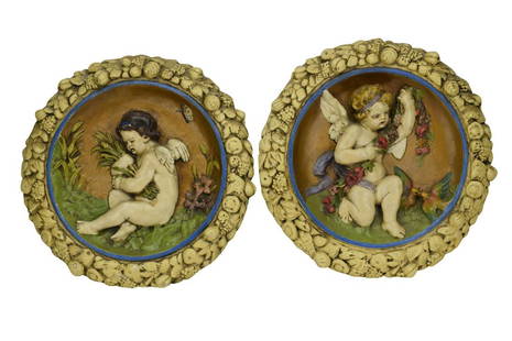 PAIR SIGNED PORCELAIN SCENIC CHERUB WALL PLAQUES: Unusual scenic round ceramic or plaster wall plaques. One with a cherub, putto, or angel in a field holding a stand of garland draped across his body, two birds at his feet, a bronze sky, blue stripe