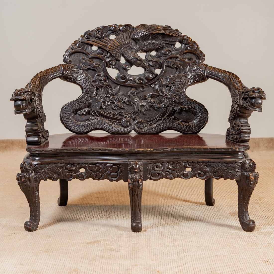 Chinese Elaborately Carved Hardwood Bench: Ebonized carved hardwood backsplat depicting a phoenix among smoke clouds, the arms extending as dragons, flat bench seat over a carved apron supported by five cabriole legs, 43 1/4 in. H, 49 1/2 in.