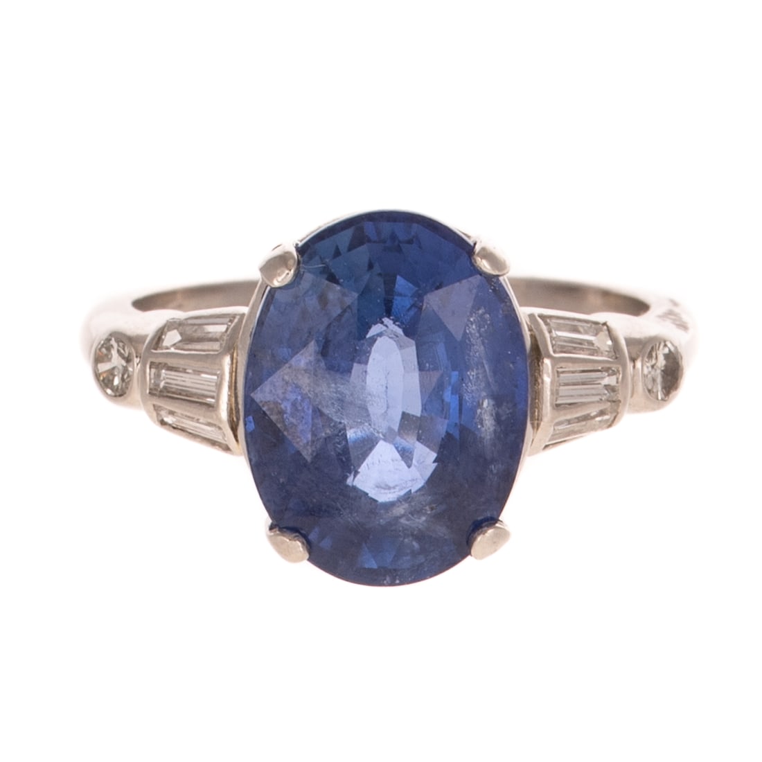 A GIA Ceylon Sapphire & Diamond Ring in Platinum: Platinum ring featuring an oval sapphire weighing 7.41 carats prong set, accompanied by GIA Gemological Report #2233722182, flanked by baguette diamonds flush set on a curve and round diamonds bezel s
