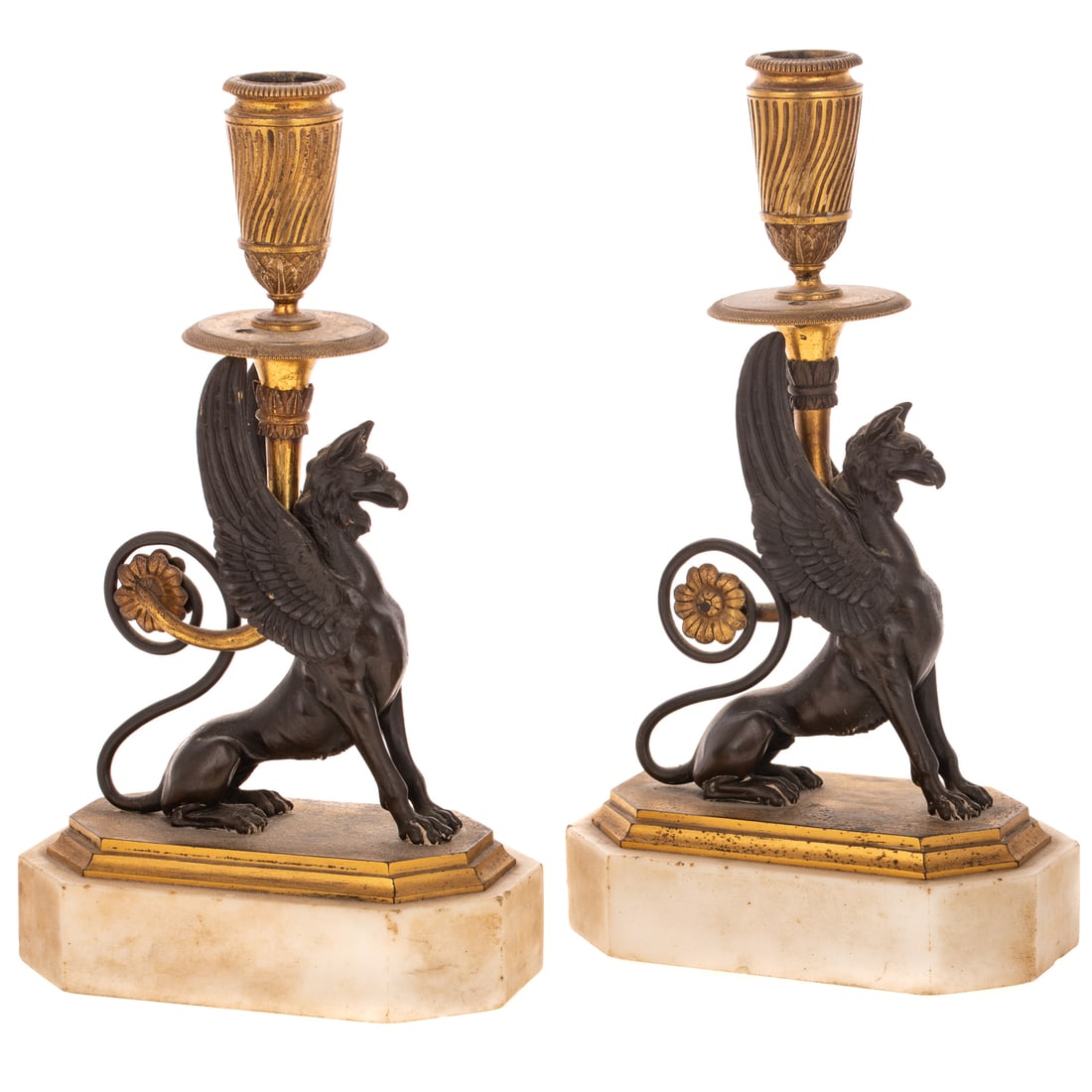 A Pair of French Bronze Griffin Candlesticks: A pair gilt and patinated bronze winged griffin candlesticks with rosette rolled up into the tail, on white marble bases, 10 1/4 in. H, 6 in. L, 3 1/2 in. W