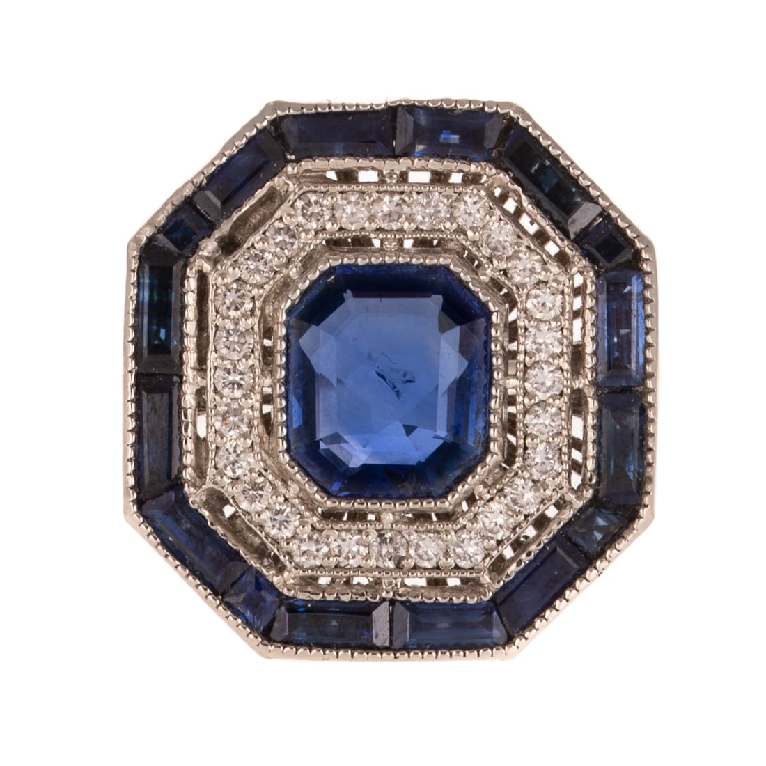 An Art Deco Style Sapphire & Diamond Ring in Platinum: Platinum ring featuring an emerald cut sapphire weighing approx. 3.04 carats bezel set in the center of a halo of round diamonds weighing approx. 0.45 ctw bead set and an outer halo of rectangular and