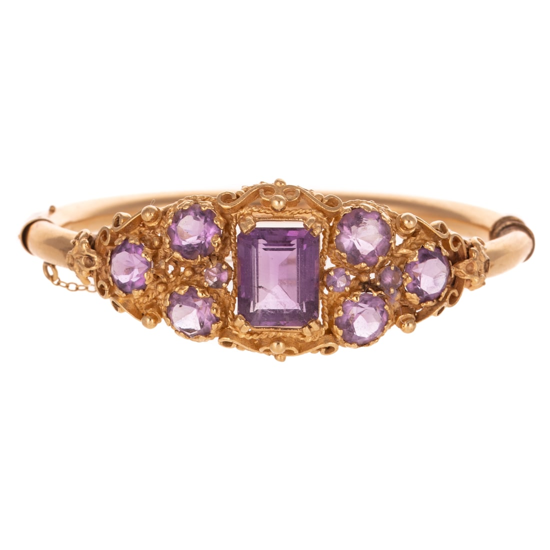 A Victorian Style Amethyst Bangle in 14K: 14K yellow gold oval hinged bangle bracelet featuring emerald and round amethyst set among a granulated scroll design on top of bangle, box clasp and safety chain, 2 1/4 interior Diam., 23.7 grams