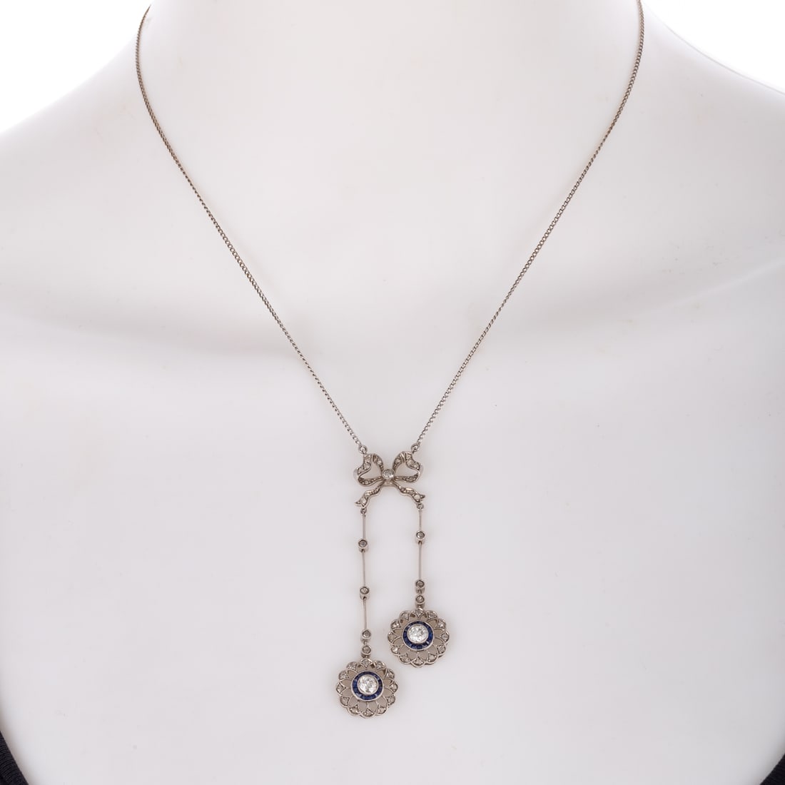 An Edwardian Diamond Negligee Necklace in Platinum: c.1900s, platinum necklace featuring diamond bead set into bow atop two asymmetrically dangling pendants with Old European cut diamonds, sapphire halo and diamond scalloped outer frame, diamonds weigh