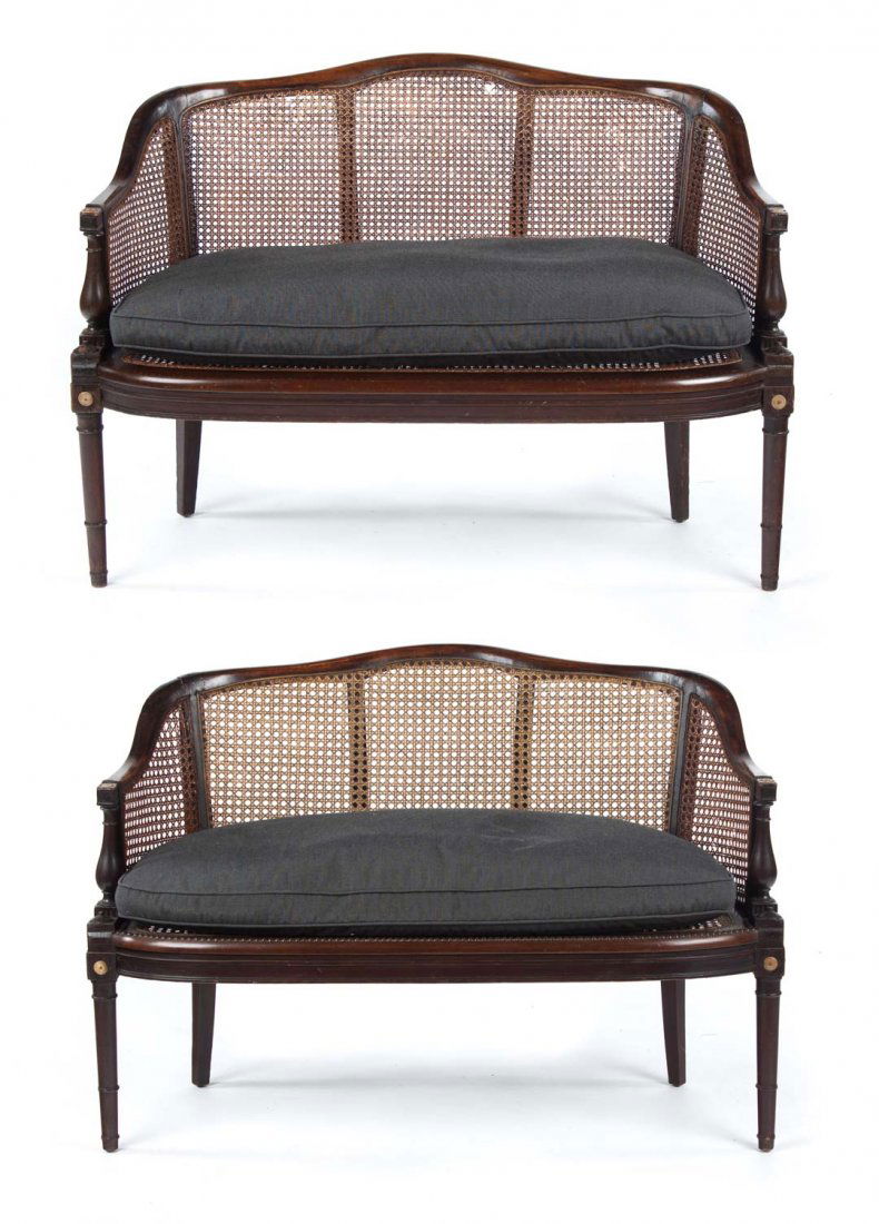 Pair of George III caned carved walnut settees: 19th century; serpentine crest rail forming sloped arms, bow-front seat rail, round-turned, and tapered legs, caned panel back, arms, and seat with upholstered loose seat cushions, carved bone roundel