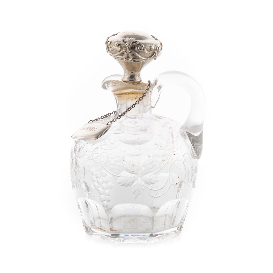 American cut glass and silver ewer: late 19th century; possibly Pairpoint, with etched grapevine and leaf decoration, silver capped stopper, includes silver-plated liquor tag, engraved "Spirits", 8 in. H.