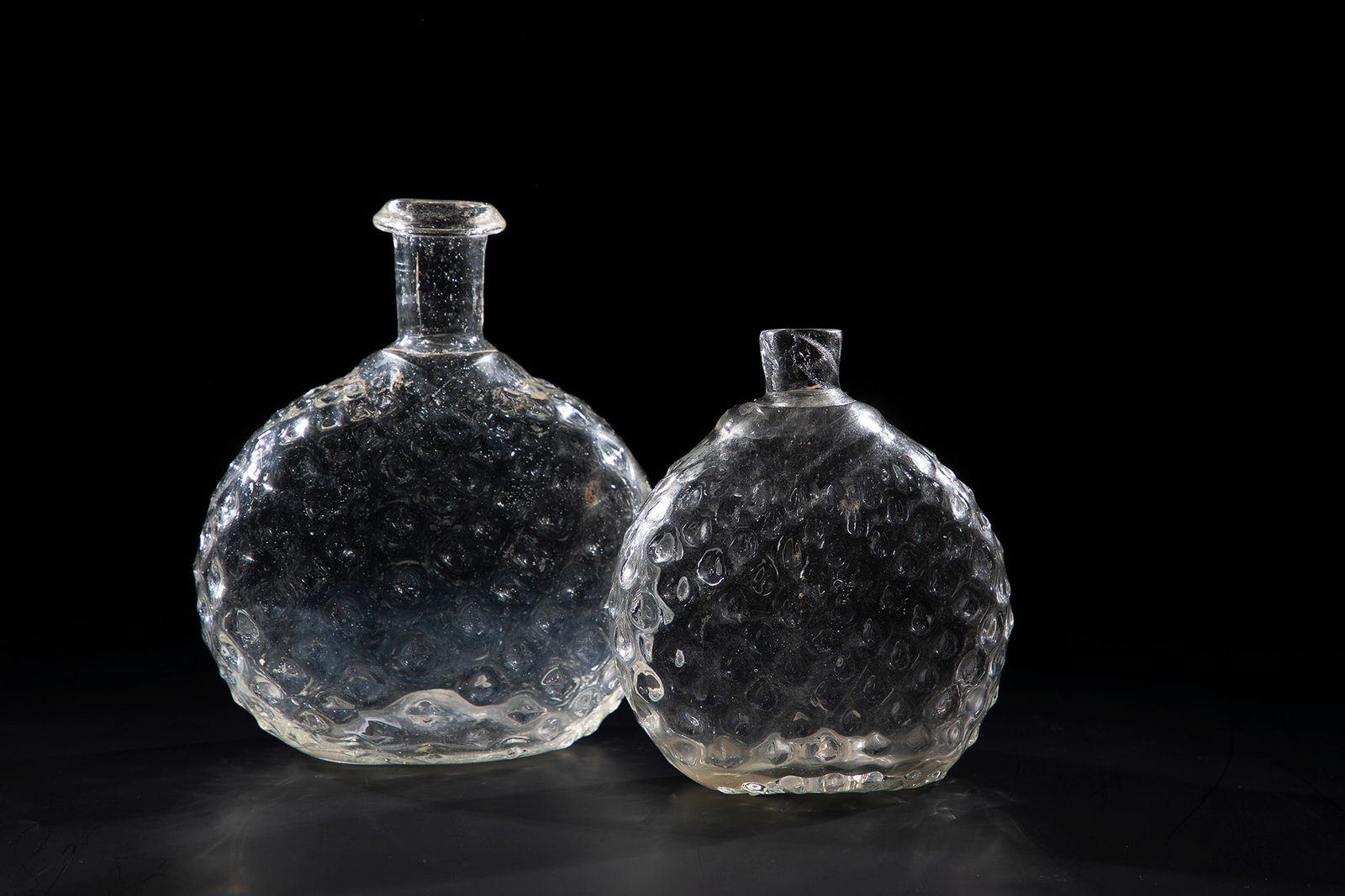 Two wart bottles: Two wart bottles, Alpine, 18th century. Grey-manganese glass with slightly raised bottom and tear-off. Spherical, double-sided flattened body with wart decoration and smooth tubular neck, one with rei