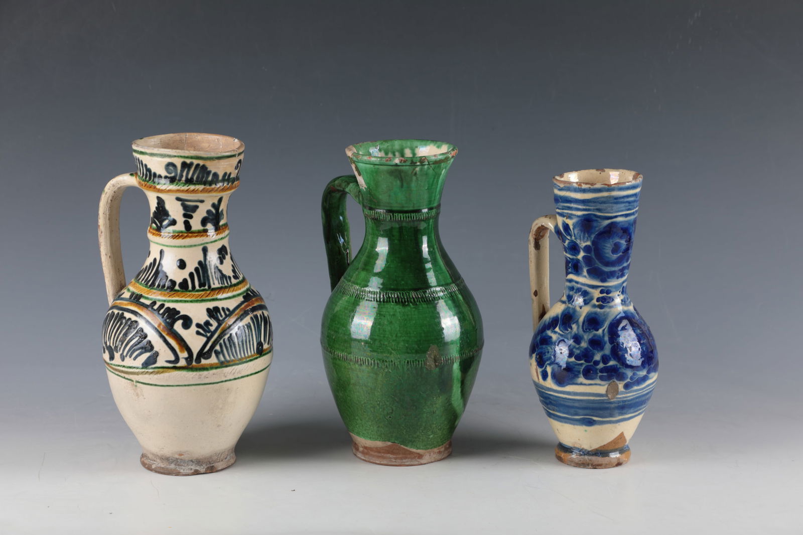Three Jugs: Three Jugs Transylvania, Kichberg, Hammersdorf, 19th century On a light background, décor in blue, green and oker or green glazed with fluted décor and rolled handle. Partly damaged. H. 22.5 to 24.5