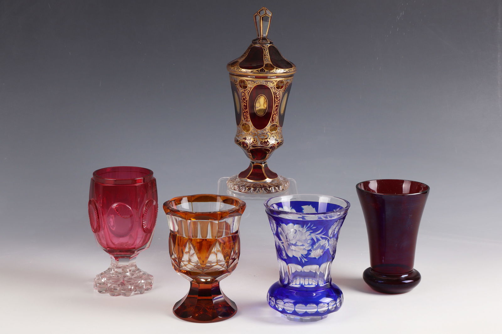 Mixed lot of four cups and a lidded goblet: Mixed lot of four goblets and a lidded goblet 19th century. Colourless glass, red glazed / blue overlay / gold ruby underlay / amber glazed. Partly decorated with cut decoration; Lidded goblet with ar