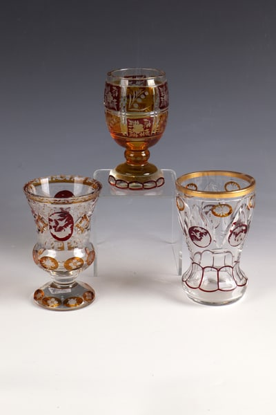 Three Cups: Three cups from the beginning of the 20th century. Colourless, partly yellow and purple glazed, cut-decorated glass with cut animal depictions and bouquets of flowers. A cup at the edge of the lips mi