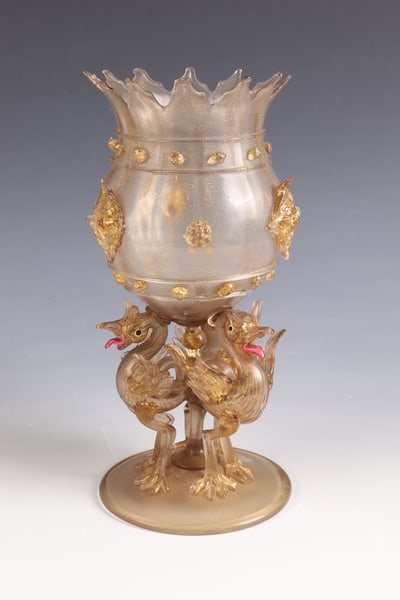 Cup: Murano goblet, c. 1900 Brown-tinted glass with gold spangles. Disc base with upturned edge and tear. Shaft of three fully three-dimensional birds holding the bulbous cuppa with rich appliqués. Serrat