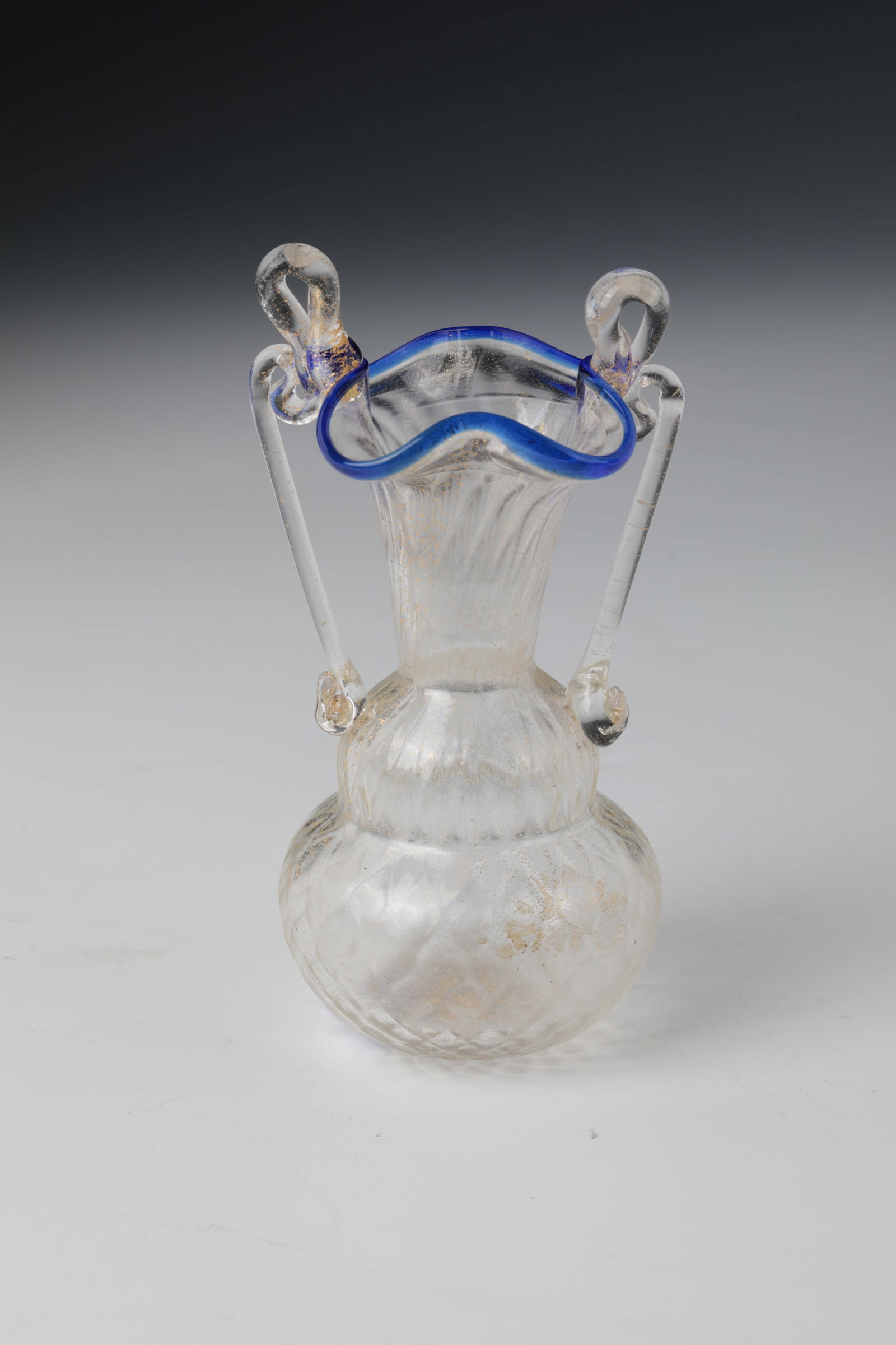 Small Handle Vase with Blue Rim: Small vase with blue rim, Murano, around 1900 Colourless glass with gold spangles. Bulbous wall with optical wrestle decoration. Trumpet-shaped widened neck with wavy muzzle rim. Appliquéd handles.