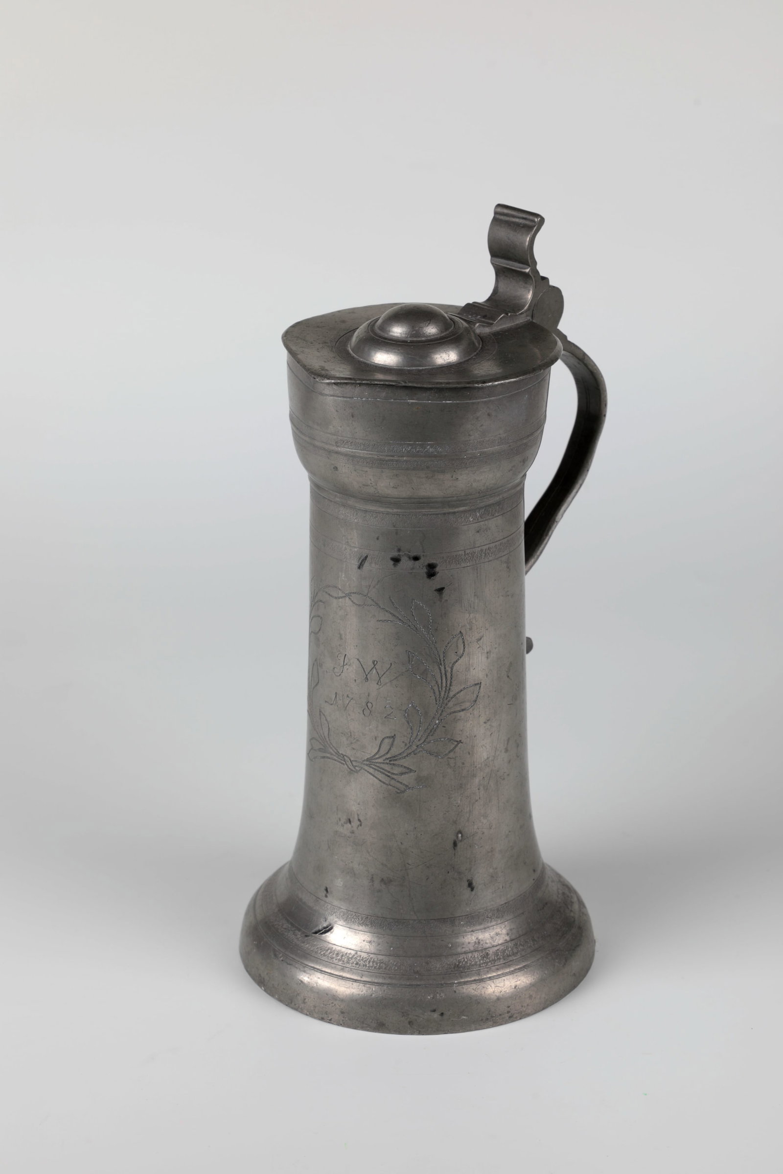 Pewter jug: Pewter jug Dated 1782 Frontal on the smooth wall initials ''JW'' and dated ''1782'' between crossed palm fronds. Tin lid with thumb rest. Ribbon handle with devious pewter mark. In the bottom flower