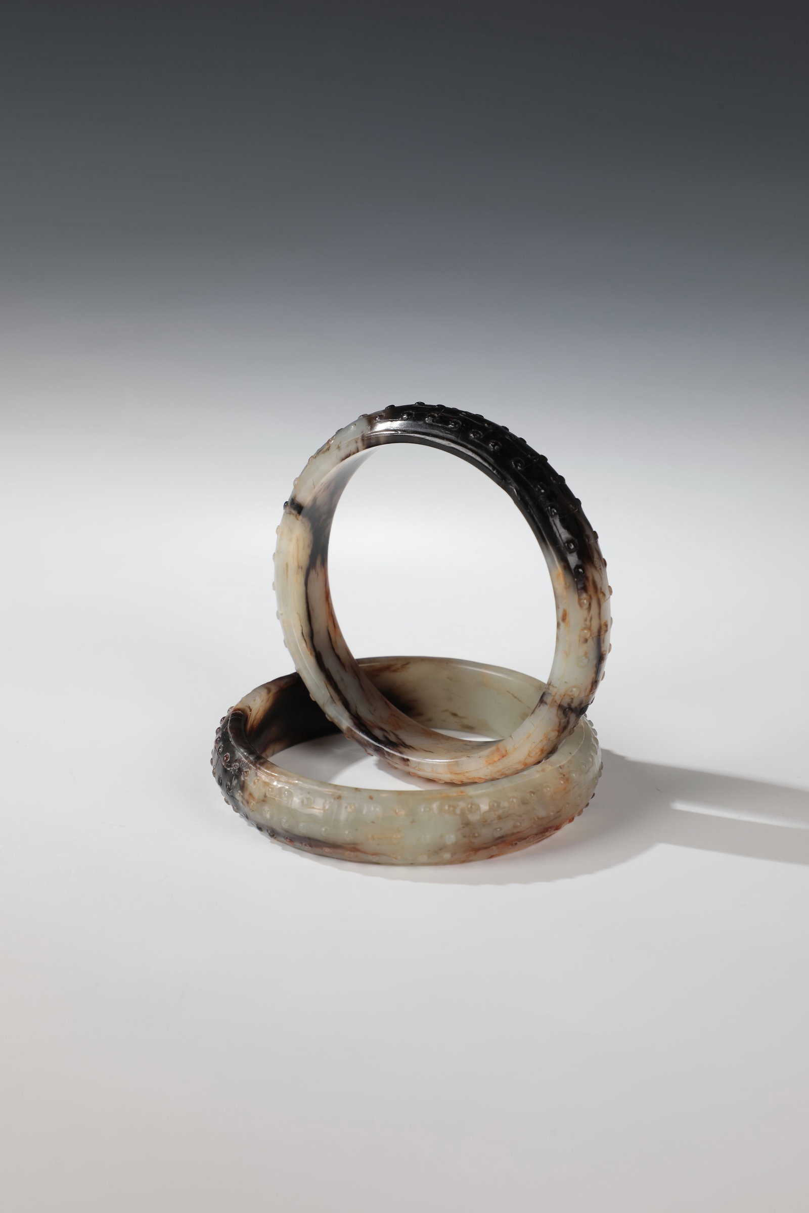 Pair of Jade Bangles: Pair of Jade Bangles in China, circa 1900 A pair of marbled jade bangles with polished outer wall and studded décor all around. Black-reddish-brown inclusions. A bangle with minor edge chips. In