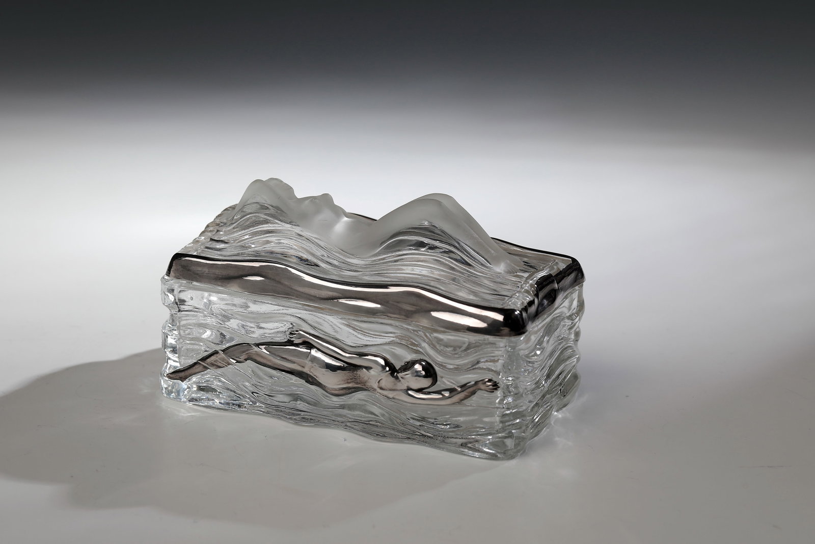 Lidded box with nude and float: Lidded box with nude and float Bohemia, 20th century Colourless glass, moulded. On the top of the lid and on the two long sides, sculpturally modelled swimmers. lid edge and two swimmers with silver