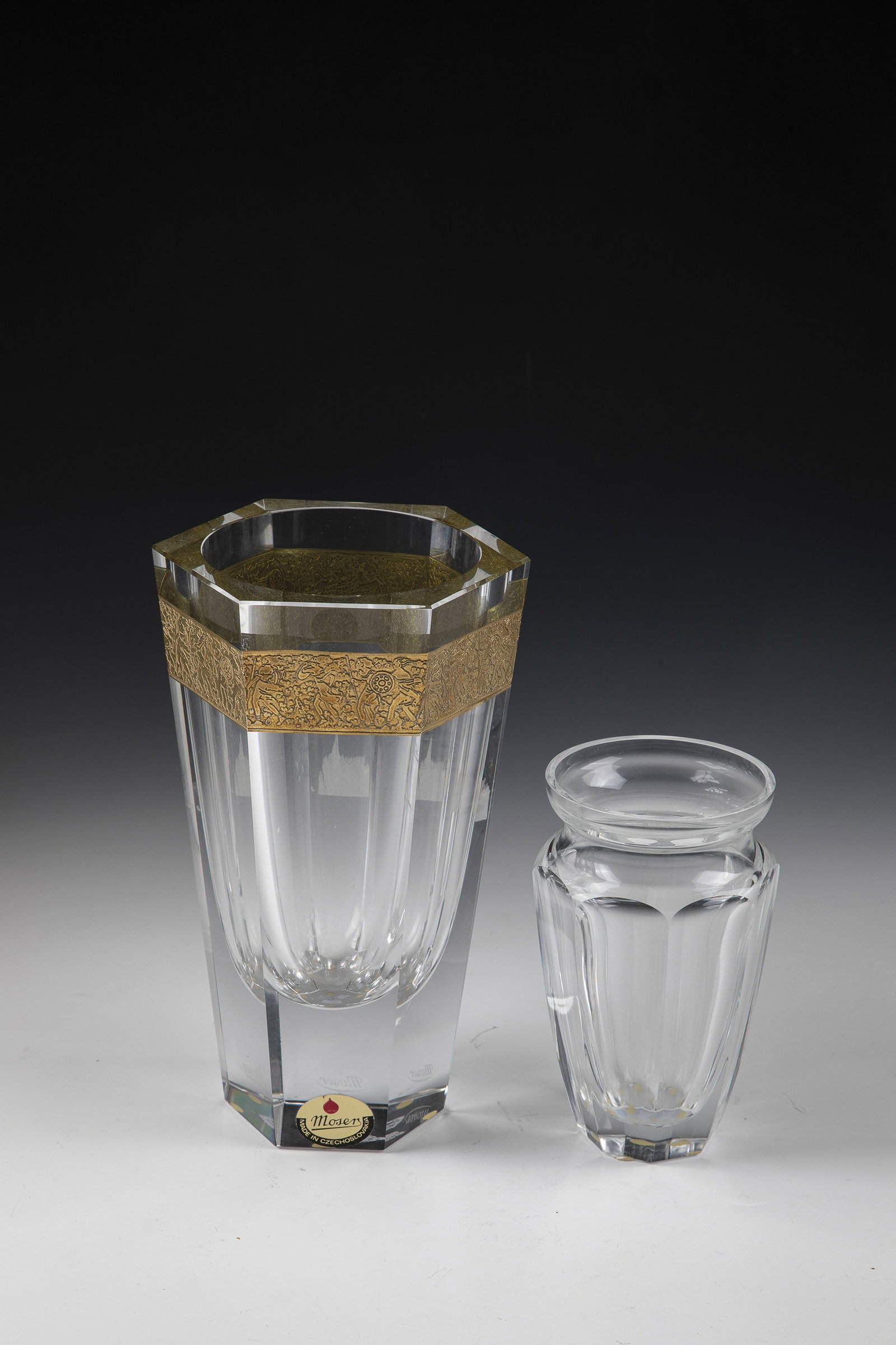 Two Vases: Two Vases Moser, Karlovy Vary, c. 1920 Colorless glass, faceted. A glass with an all-round relief-etched, gilded and brown patinated frieze with Amazons. Both vases with etched stamp ''Moser''. H.